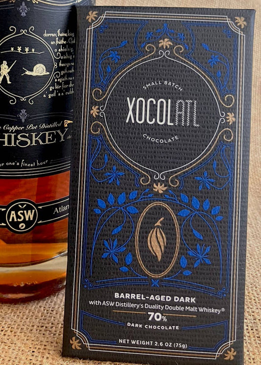 A bar of XocolAtl Dark Chocolate next to a bottle of ASW Duality Double Malt Whiskey, both with branding and flavor information on the packaging.